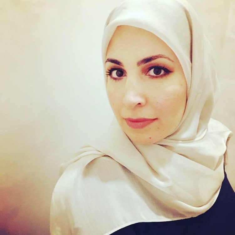 Mona Haidar smiles in front of a white wall, she wears a white scarf around her head.