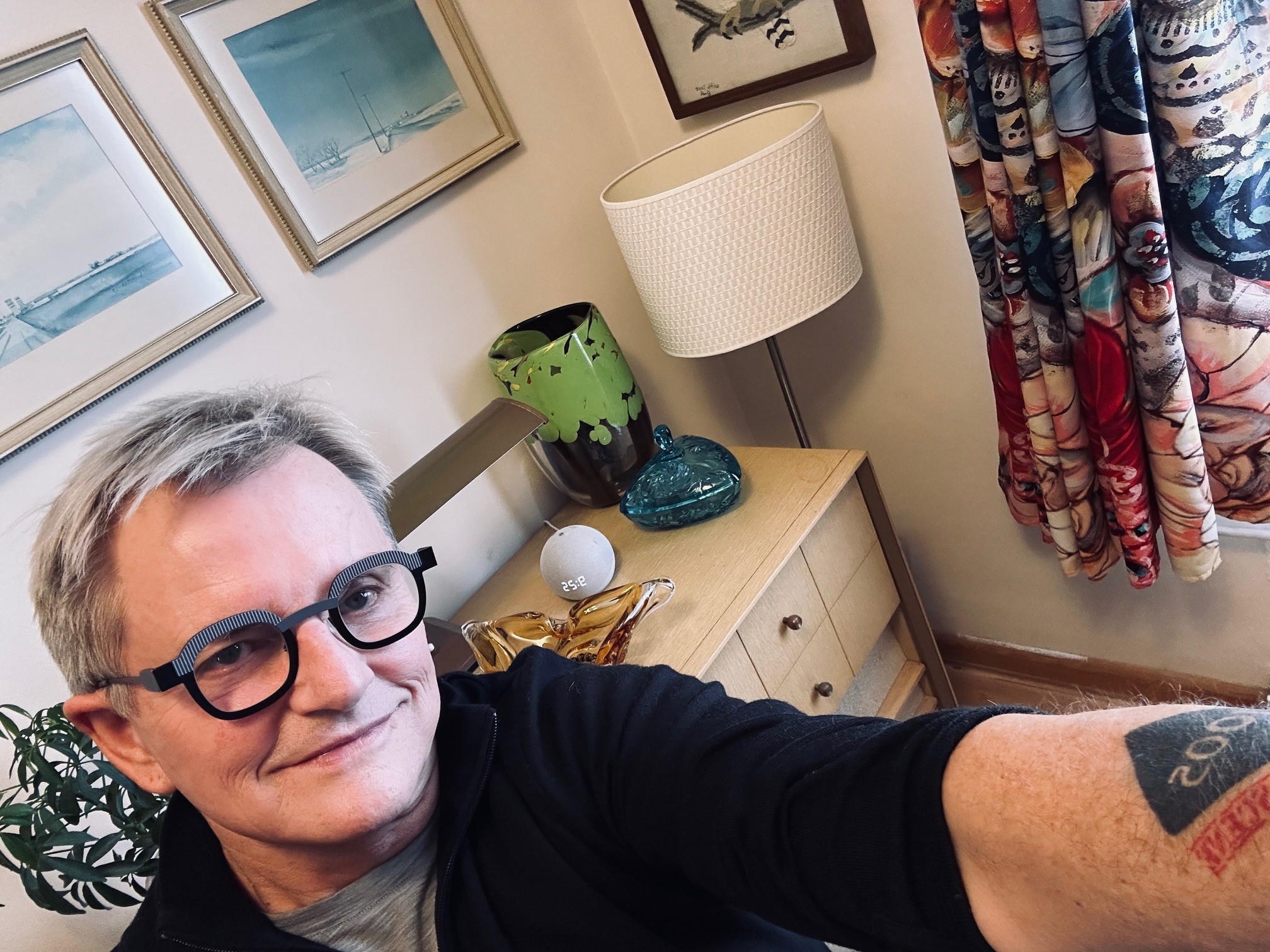 Dennis Sumara smiles wearing glasses with a vase, lamp, and dresser visible in the background.