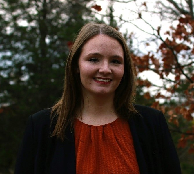 White woman with dark hair smiles at the camera wearing a red shirt and black cardigan white standing outside with fall leaves in the background