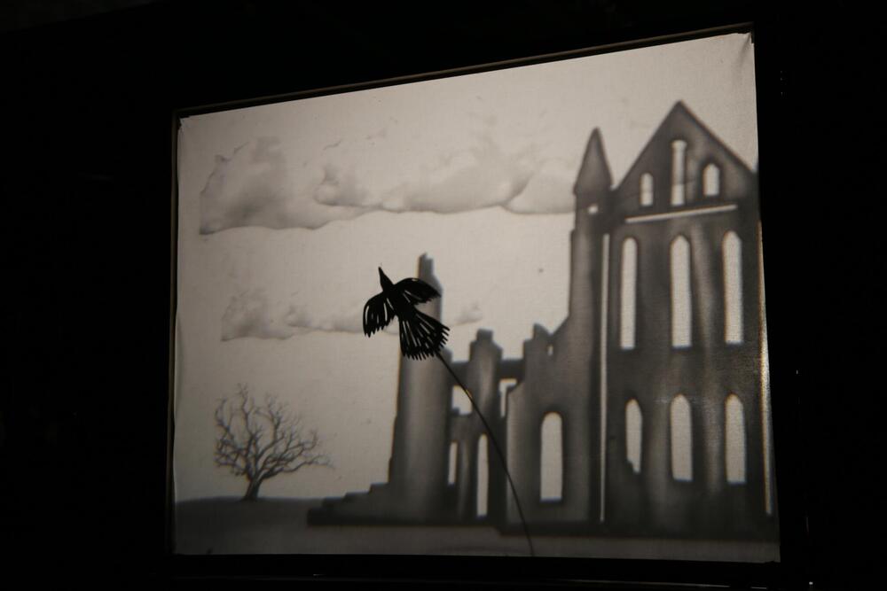 A shadow puppet screen displays a bird flying in front of a monastery.