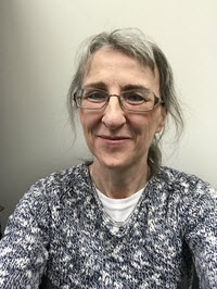 Brenda Reed smiles wearing glasses and a floral shirt against a white wall.