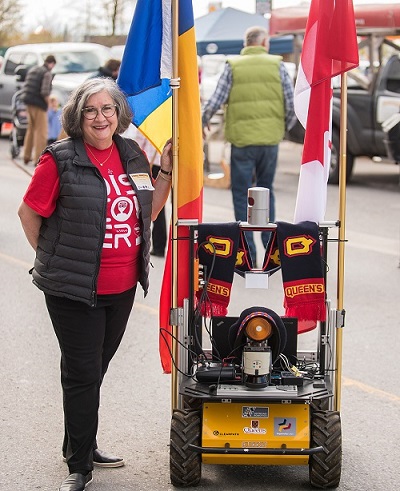 Lynda Colgan poses outside next to a rolling robot that holds a Queen's flag.