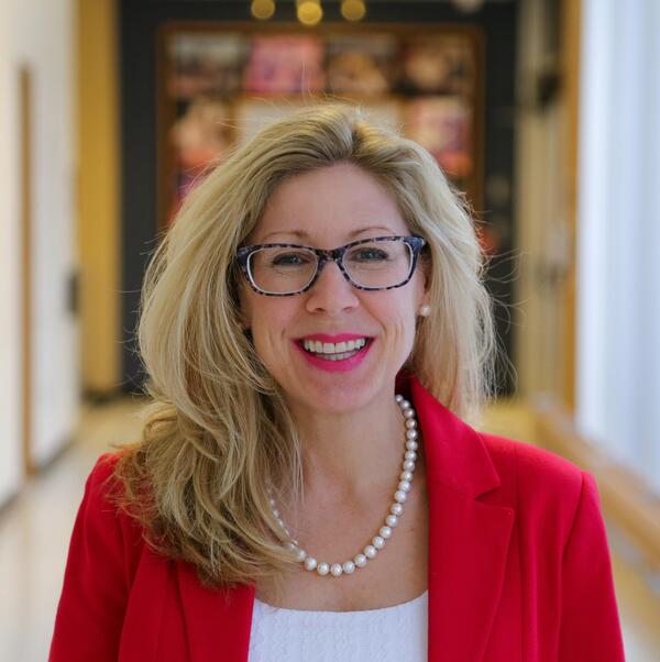 Christine Armstrong smiles in a hallway wearing glasses and a bright red blazer.