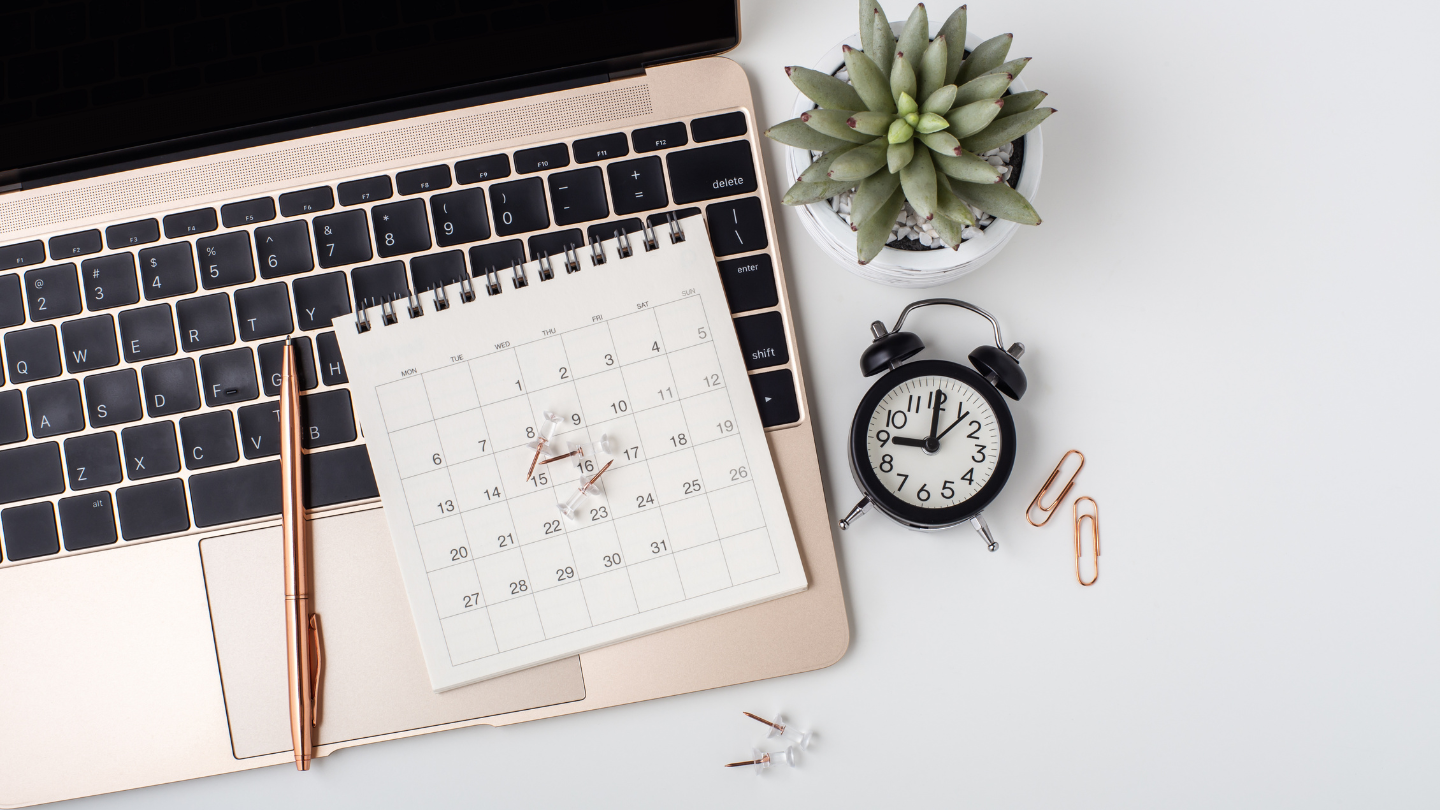 A laptop in a workstation with a calendar, clock, and plant