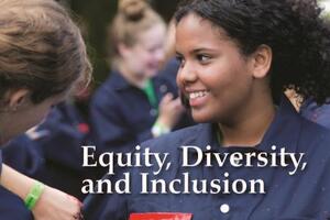 A student smiles as she is having her coveralls painted in Queen's Tri-colour with words "The Knowledge Forum, Equity, Diversity, and Inclusion"