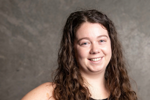 Dayle stands with one hand on hip and smiles. The background is grey.