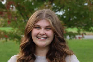 Hanna is smiling at the camera, standing outside with a tree and green grass in the background