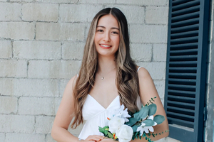 Mya smiles at the camera wearing a white dress and holding flowers. She stands in front of a brick wall