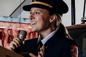 Kristy-Lynn smiles while holding a microphone and gesticulating. She is wearing a uniform. 