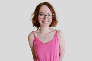 Claire, wearing a pink dress is smiling at the camera. She has red, chin length hair and is wearing glasses