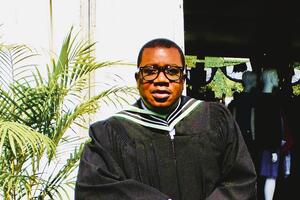 Dr. Baker looks at the camera wearing glasses next to a fern plant. He is wearing a graduation gown and hood.