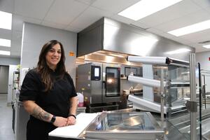 Grace smiles at the camera in an industrial kitchen that is full of stainless steel appliances and surfaces