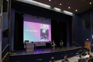 Lee Airton speaks on a stage with a large graphic that reads "Lee Airton" on a projected screen.