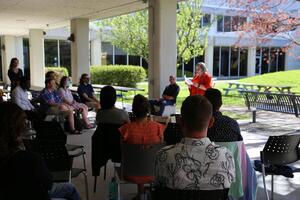 People sit in a semi-circle outdoors on a sunny day while Rebecca Luce-Kapler speaks.