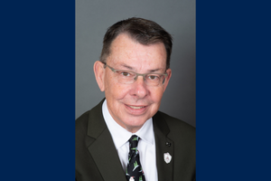 Brian Evoy smiles wearing glasses and a suit and tie. Brian also wears a pin on his lapel.