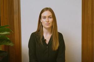 Alex Lowes sits in front of a white wall. There is a plant to her left.