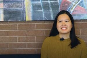 Jenny Lee Northey smiles in front of a brick wall with a painting hanging on it.