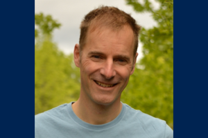 David Rankine smiles outside wearing a light blue t-shirt in front of green trees.