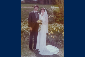 John and Donna Greenhorn smile in a wedding photo outside in front of flowers in a garden. They wear wedding attire and Donna holds a bouquet of flowers.