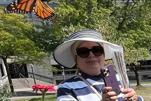 Maha Al Makhamreh smiles wearing a hat and sunglasses in a garden and holding a cellphone. There is a butterfly flying in the foreground.