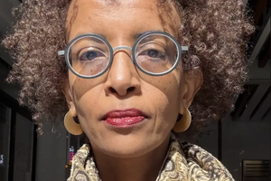 Tigist Amdemichael smiles wearing glasses, red lipstick, and scarf and poses in front of the light from a window.