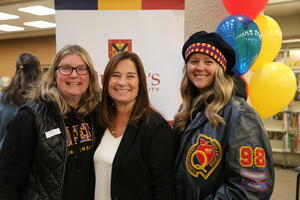 Three alumni stand in front of a banner and balloons. The woman on the right wears a tam and a Queen's leather jacket.