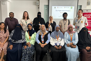 A group of educators in burkhas and headscarves smile with instructors at a certificate ceremony. A screen in the back reads "The Night School Project."