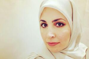 Mona Haidar smiles in front of a white wall, she wears a white scarf on her head.