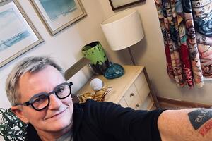 A man with grey hair taking a selfie with a tatoo on his arm and stylish black glasses on