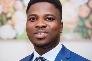 A black man wearing a blue suit and tie smiles at the camera