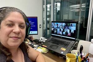 A woman looks at the camera with a computer next to her with online students engaging in a learning activity