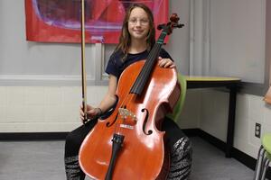 A student poses with cello in a classroom while sitting on a chair.