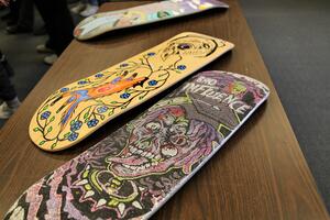 Two skateboard decks sit on a table. One is painted and one has a design of a face made our of various glittery paints and materials.