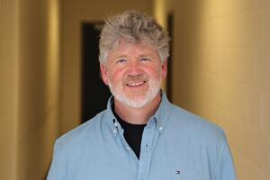 Man with grey hair standing in a hallway, wearing a blue button down shirt