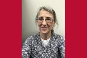 Brenda Reed smiles wearing a floral shirt and glasses. The photo is against a red background.