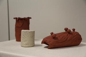 Three clay artworks on a plinth. Two are terracotta clay and one is a small white clay cup.