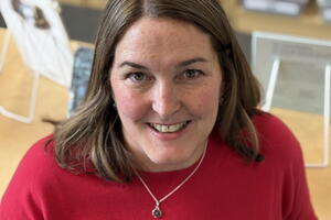 Tracey Mailloux smiles wearing a red shirt and necklace and she stands in front of shelves of classroom materials.