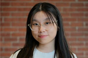 Wennie Chen Smiles wearing a white shirt and white button overshirt t-shirt and glasses. Her hair is down around her shoulders and she stands in front of a red brick wall.