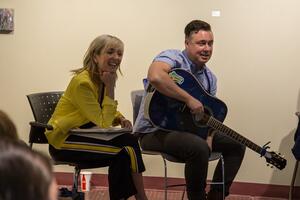 Taes Leavitt and Nick Adams sit engaged in chairs. Nick holds a guitar across his lap.