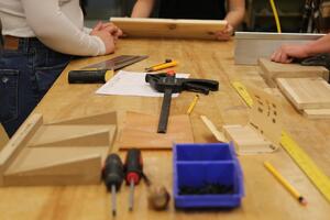Tools sit on a wood workbench. A student uses a saw to cut some wood.