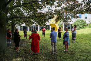 A group stands in a circle outside with a yellow sculpture in the background.