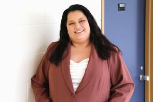 A woman with dark hair smiles at a camera wearing a white shirt and a brown blazer
