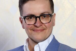 Lee Airton smiles wearing glasses, a blue suit jacket, and a white collared shirt.