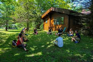 People sit in a circle on the grass in front of a log cabin.