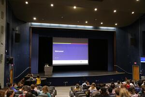 Students sit in an auditorium with a large screen at the front that reads "Welcome."