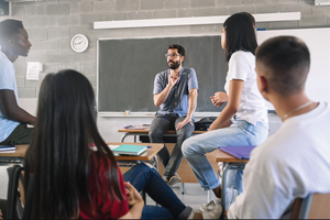 A teacher sits on a table speaking to four adult students who sit at desks.