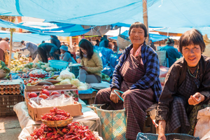 Two women sit at a market with bags and baskets of food.