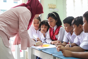 A teacher in a headscarf helps students at desks.