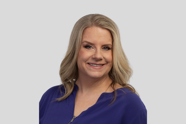 Amy Burns smiles at the camera. She has blonde hair and is wearing a deep purple blouse.