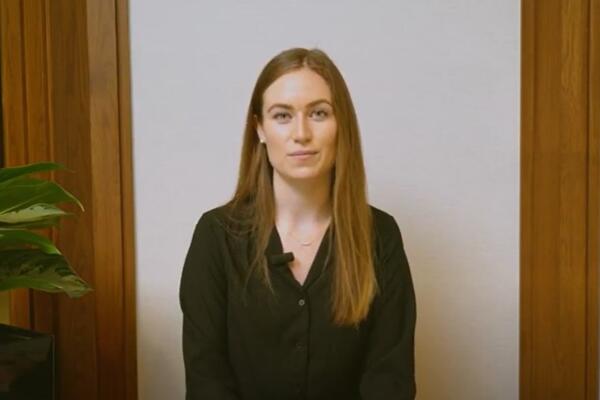 Alex Lowes smiles in front of a white wall. There is a plant to her left.